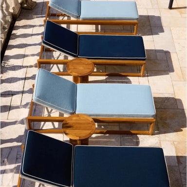 Four wooden chaise lounge chairs with light and dark blue cushions arranged in a row, with two small round wooden side tables on a tiled patio.