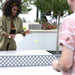 A woman in an olive green shirt playing ping pong with a yellow ball, with another person in a pink shirt partially visible in the foreground on an outdoor patio.