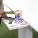 A white outdoor table with a built-in tray holding a La Croix sparkling drink can, with a person's hand resting on the table edge and blurred green grass in the background.