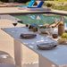 A white outdoor dining table set with plates, wine glasses, a bottle, and food near a swimming pool in a sunny patio setting.