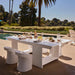 A modern white outdoor dining table set with plates, glasses, and a bottle, accompanied by matching white curved chairs, situated next to a swimming pool with palm trees and a landscaped garden in the background.