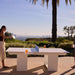 A white outdoor ping-pong table set on a patio with a woman on the left holding a paddle and a person partially visible on the right, with a palm tree and distant city skyline in the background.