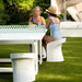 A woman in a blue patterned swimsuit and straw hat sits on a white curved outdoor chair at a white table on a green lawn, with another woman seated across from her in a patio setting.