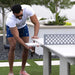 A man in a white t-shirt and blue palm tree print shorts leans over a white outdoor table, holding a beer bottle in a patio setting with green grass and plants in the background.