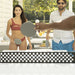 A lifestyle shot of a ping-pong game being played on an outdoor patio. A woman in a red bikini and a man in a white striped shirt are standing behind a table with ping-pong paddles, while a person's arm in the foreground holds a paddle.