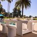 A modern outdoor dining set with a white rectangular table and matching curved white stools, styled on a patio beside a swimming pool with palm trees and a blue umbrella in the background.