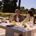A man and a woman sit at a white outdoor dining table set with wine glasses, a bottle of rosé, cheese, grapes, and salads, in a sunny garden environment.