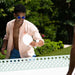 A man outdoors wearing a light peach button-up shirt, blue shorts, and blue-tinted sunglasses, holding a white paddle in front of a green garden background.