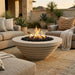 A round concrete fire pit table with a glowing fire in the center, set on a stone patio between two beige outdoor sofas with pillows and a throw blanket.