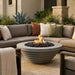 A round concrete fire pit table with black stones and visible flames sits in front of a sectional outdoor sofa on a stone patio. The scene includes beige, taupe, white, and rust pillows, plants, and a house exterior in the background.