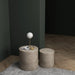 Two gray cylindrical concrete outdoor side tables on a striped rug, one with a lamp, newspapers, and a small jar, against dark green walls and wooden floor