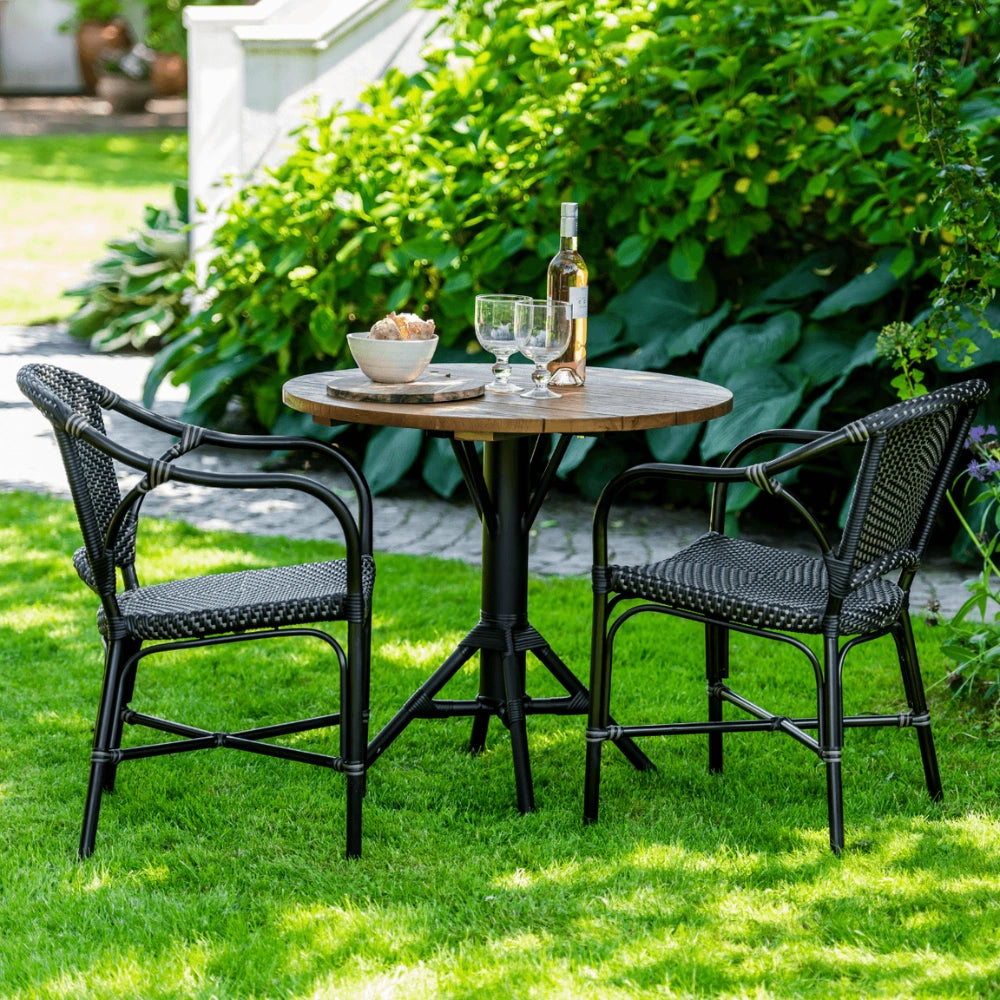 Black rattan outdoor dining armchair beside a round teak café table with a black pedestal base.