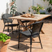 Black rattan outdoor dining armchair beside a teak dining table on a sunny patio with a woman in the background.