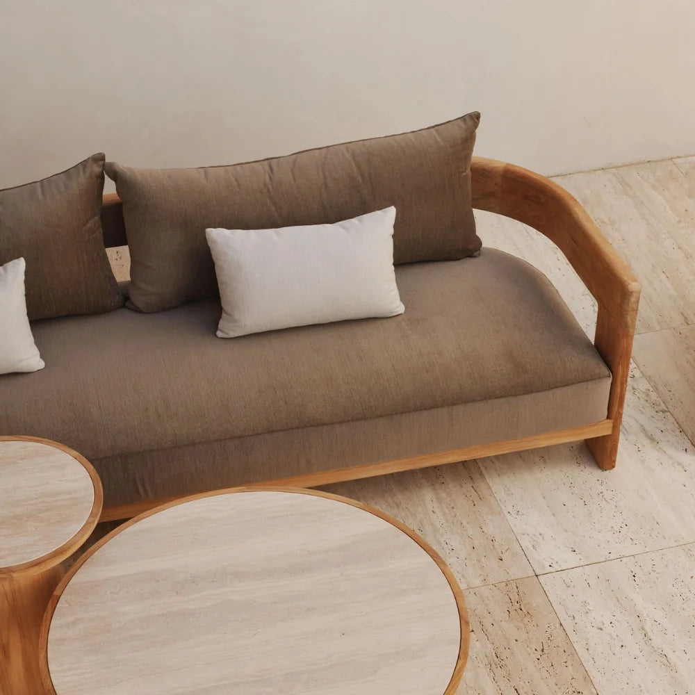 Wooden outdoor 3-seater sofa with brown cushions, white pillows, and two beige round textured coffee tables on tiled patio.
