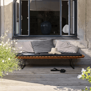 Wooden slatted outdoor lounger with black cushion set on a wooden deck patio beside a large window