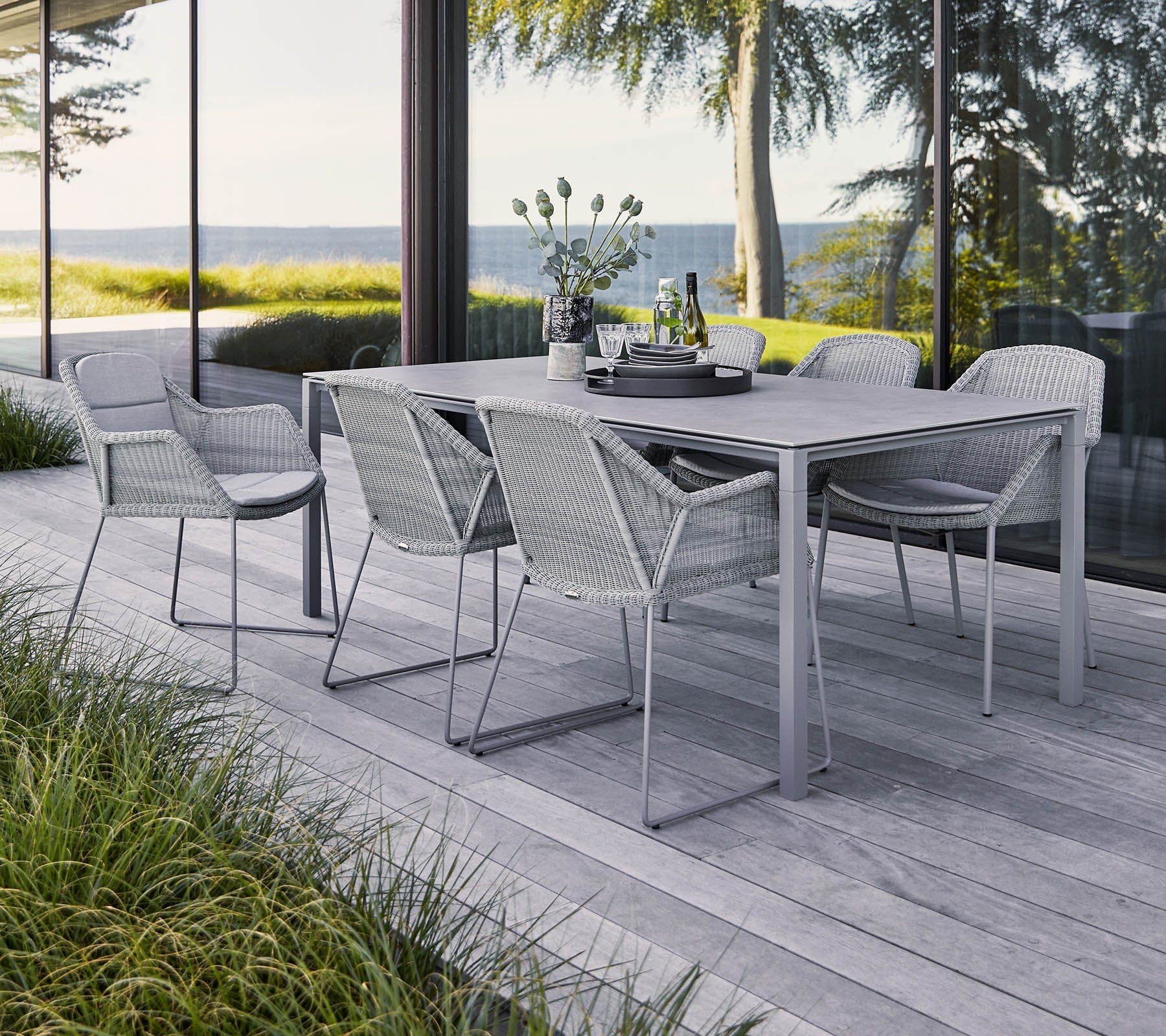 Boxhill's Breeze Dining Weave Chair Light Grey lifestyle image with dining table on wooden platform beside glass wall