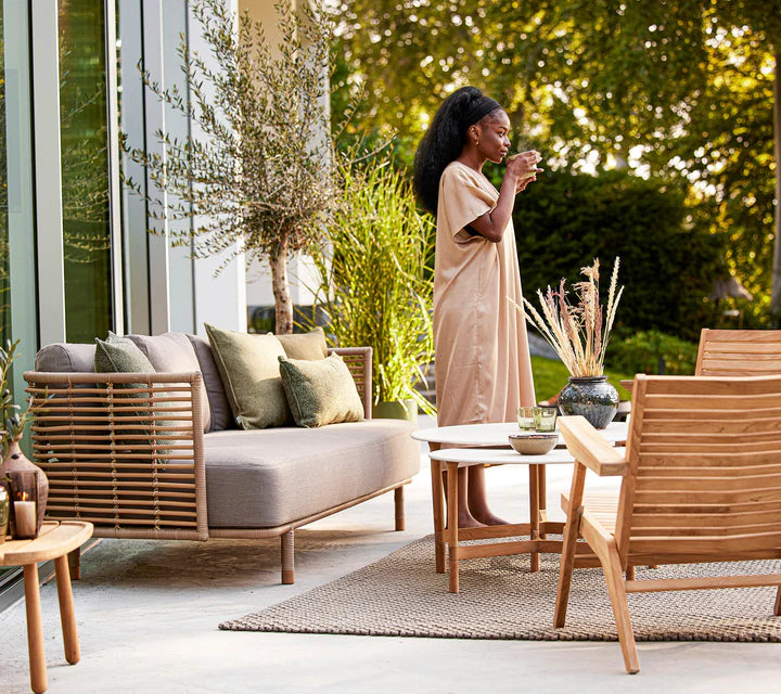 Boxhill's Sense light brown 3-seater outdoor sofa with teak chair and 2 white round tables and a woman standing drinking a coffee