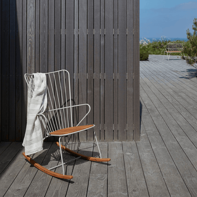 A white metal outdoor rocking chair with bamboo lamellas seat placed on a wooden deck beside wooden wall