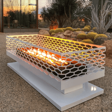 White outdoor rectangular fire pit with geometric cut-out design with flames inside placed on an outdoor patio near desert landscape with cactus plants