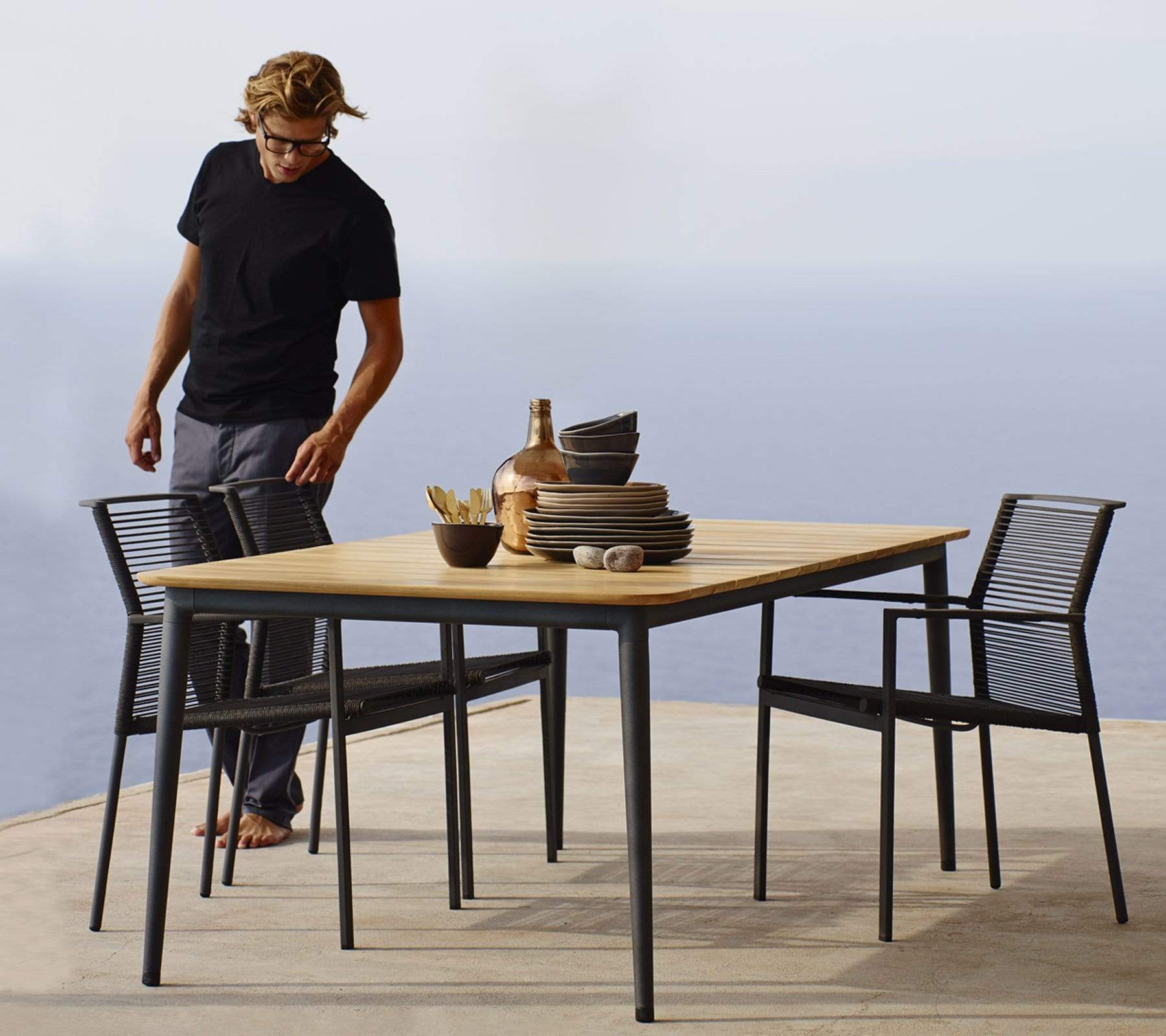 Boxhill's Edge Stackable Outdoor Armchair lifestyle image at seafront with teak top dining table and a man standing at the side