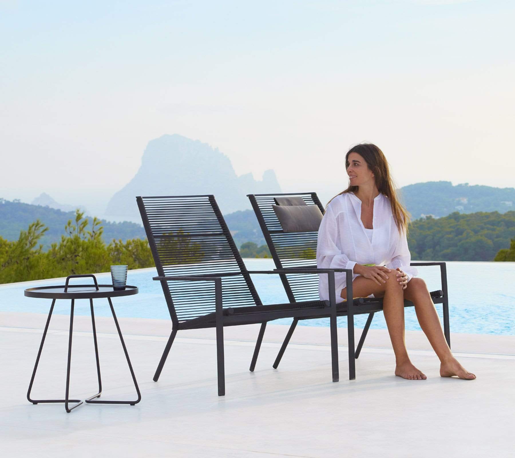 Boxhill's Edge Stackable Pool Side Highback Chair lifestyle image beside the pool with round side table and a woman sitting down 