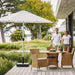 Boxhill's Hampsted Outdoor Dining Armchair lifestyle image at patio with teak round dining table and a parasol at the side, and 2 people walking at the side