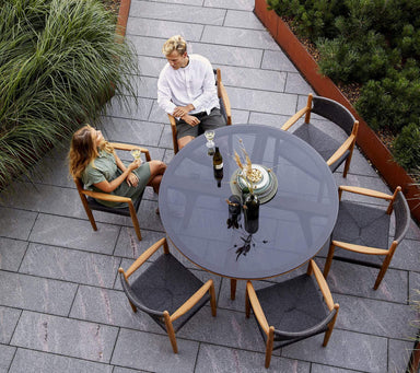 Boxhill's Aspect Teak Round Dining Table Clear Glass | Smokey Black lifestyle image with dining chairs and 2 people sitting down