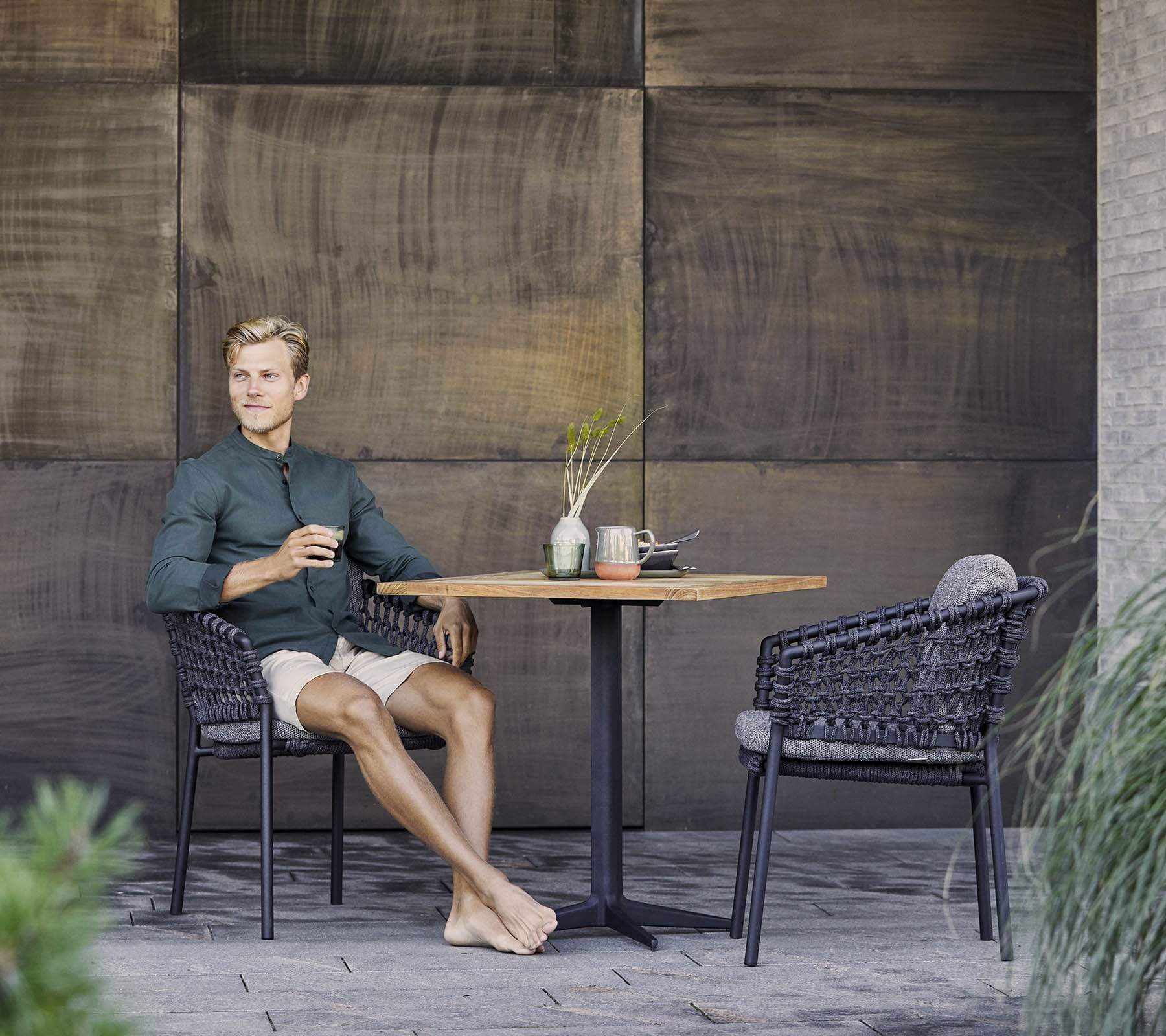 Boxhill's Drop Square Outdoor Cafe Table Lava Grey Base Teak Top lifestyle image with 2 dining chairs and a man sitting down holding a glass of coffee