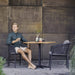 Boxhill's Drop Square Outdoor Cafe Table Lava Grey Base Teak Top lifestyle image with 2 dining chairs and a man sitting down holding a glass of coffee