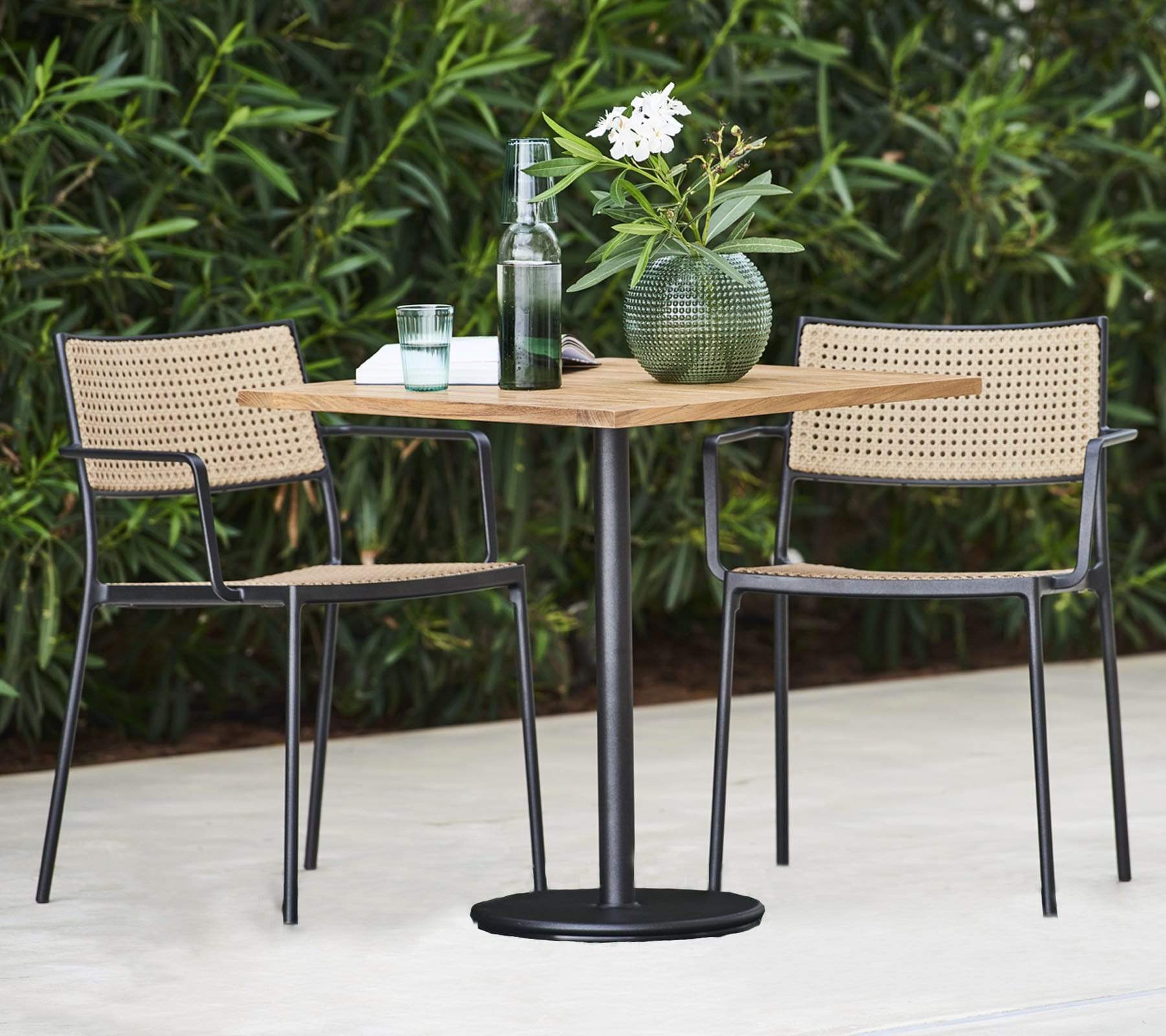 Boxhill's Go Outdoor Square Teak Café Table lifestyle image wih glass of water, bottle water container and a plant in a round vase on top, and 2 dining chairs near the plants at patio