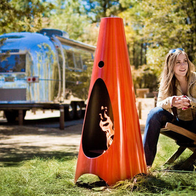 Orange cone-shaped outdoor metal fire pit stand on a grassy area beside of a woman sitting on a wooden chair