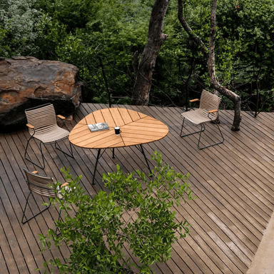 A Leaf shaped wooden outdoor dining table and 3 beige outdoor dining chairs set on a wooden deck surrounded by trees