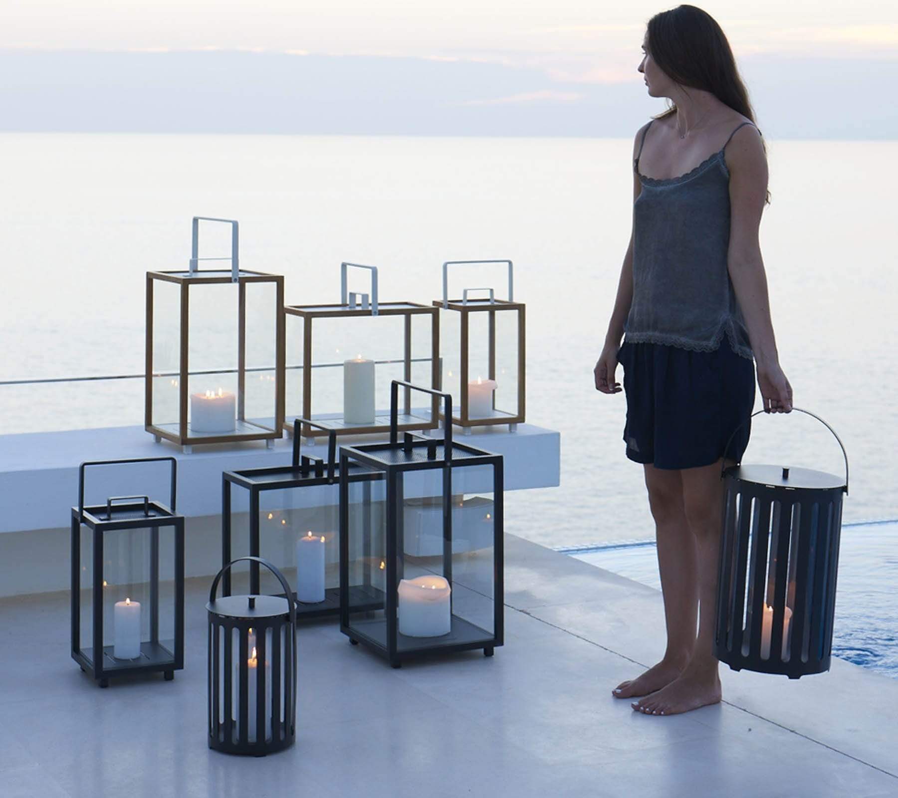 Boxhill's Lighttube Outdoor Large Lantern | Set of 2 lifestyle image with Lighthouse Outdoor Large Aluminum Lantern and Lighthouse Outdoor Large Teak Lantern with a woman standing at the side carrying Lighttube Outdoor Large Lantern