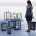 Boxhill's Lighttube Outdoor Large Lantern | Set of 2 lifestyle image with Lighthouse Outdoor Large Aluminum Lantern and Lighthouse Outdoor Large Teak Lantern with a woman standing at the side carrying Lighttube Outdoor Large Lantern