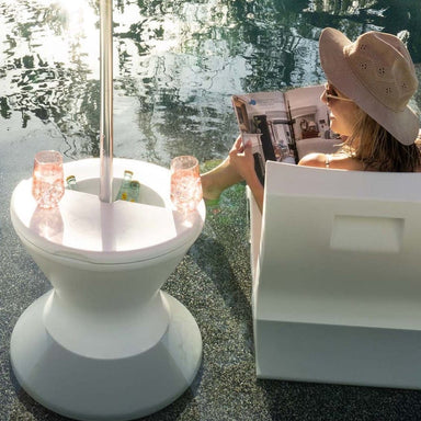  white tall ice bin side table placed in a pool beside a woman sitting on a chair reading a magazine