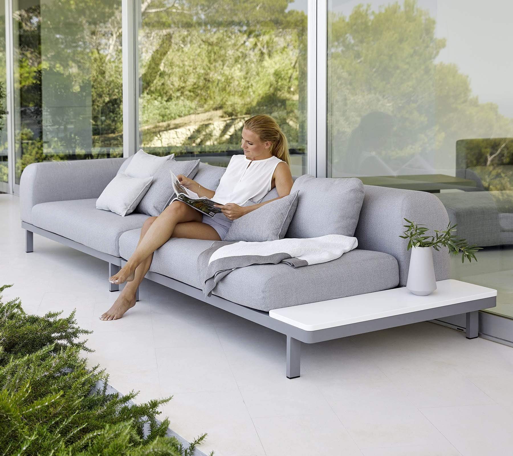 Boxhill's Space light grey outdoor sectional sofa placed against glass wall with a woman sitting on it reading a magazine