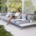 Boxhill's Space light grey outdoor sectional sofa placed against glass wall with a woman sitting on it reading a magazine